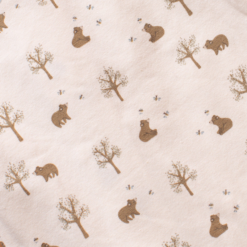 A close-up of a light cream fabric printed with a repeating pattern of brown bears, trees, and small bees.