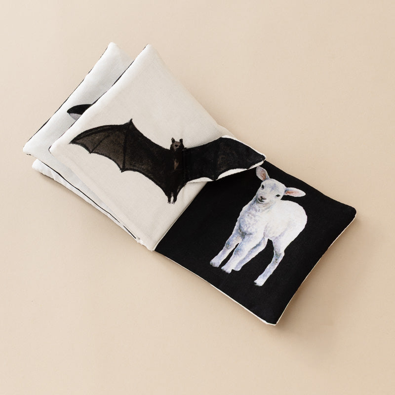 A folded fabric book accessory with a black-and-white split design, featuring a printed bat on the white side and a white lamb on the black side, photographed on a plain beige background.