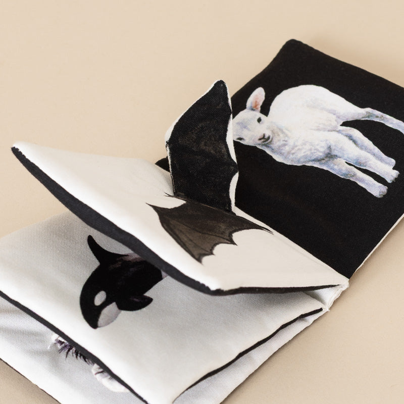A folded fabric book with black and white pages, featuring printed animal illustrations including a white lamb and a black-and-white orca, photographed on a plain beige background.