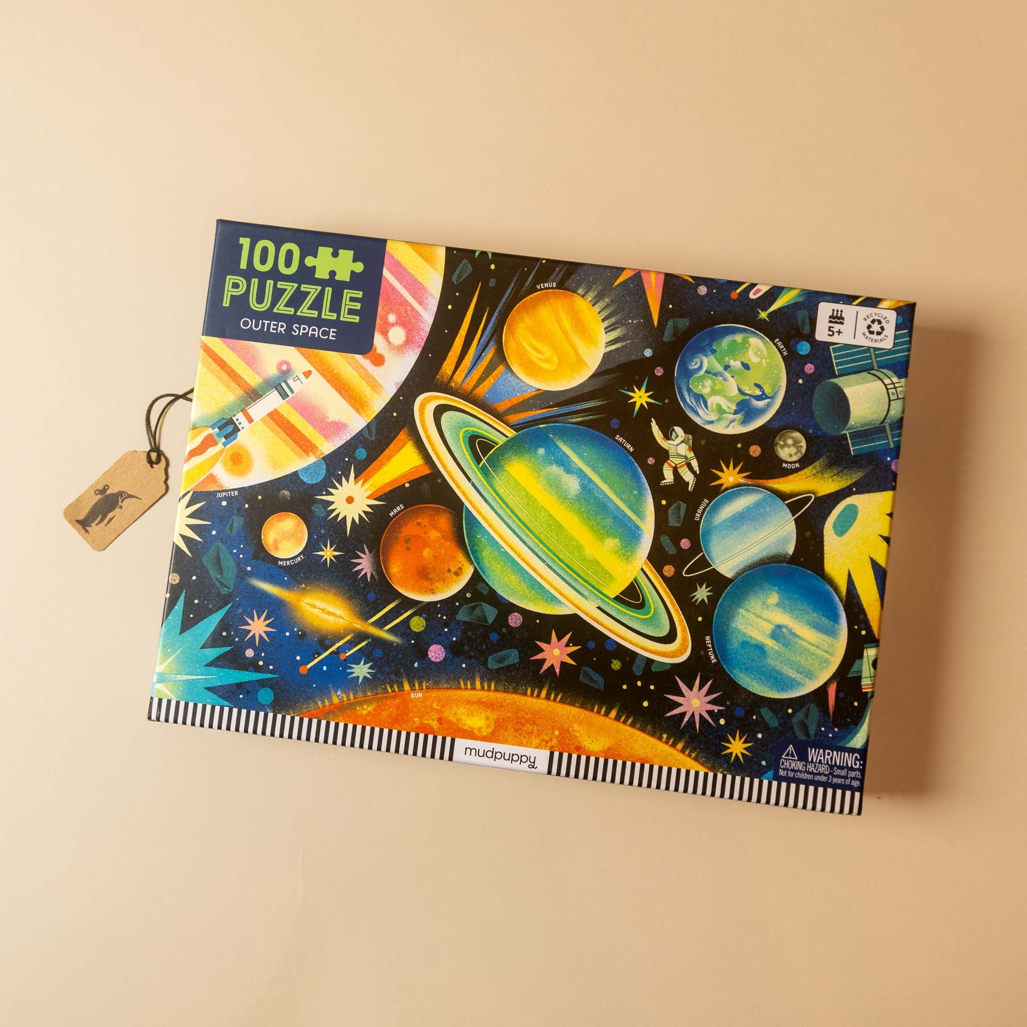 A 100-piece outer space puzzle box featuring a colorful illustration of planets, stars, and a rocket on a black background, shown on a beige surface with a small hanging tag.