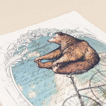 Load image into Gallery viewer, detail-image-of-bear-holding-his-feet-atop-a-tricycle-framed-by-blue-skies-and-scrolling