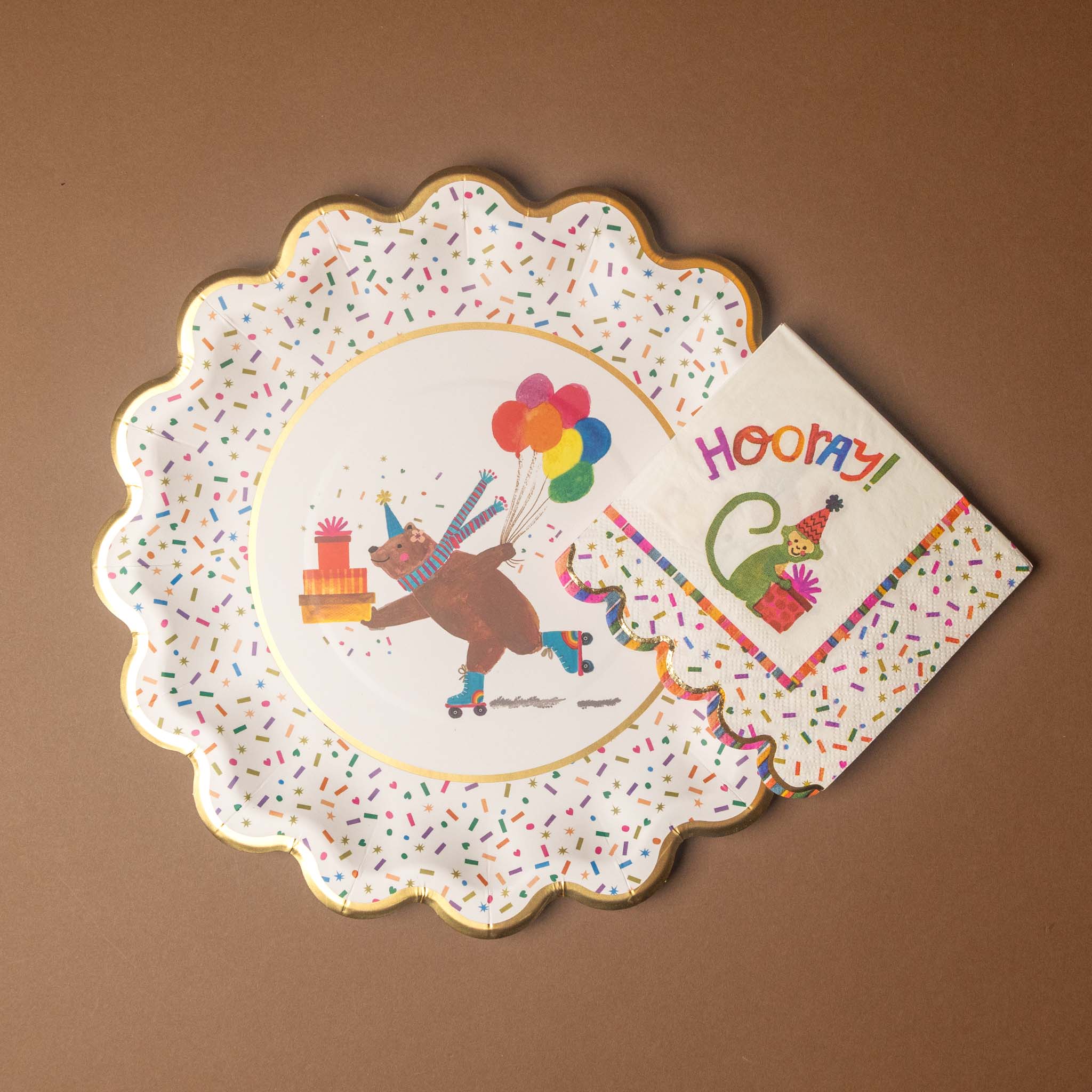 A scalloped paper party plate with a confetti speckled border and a cartoon bear holding balloons, shown with a matching napkin on a brown background.