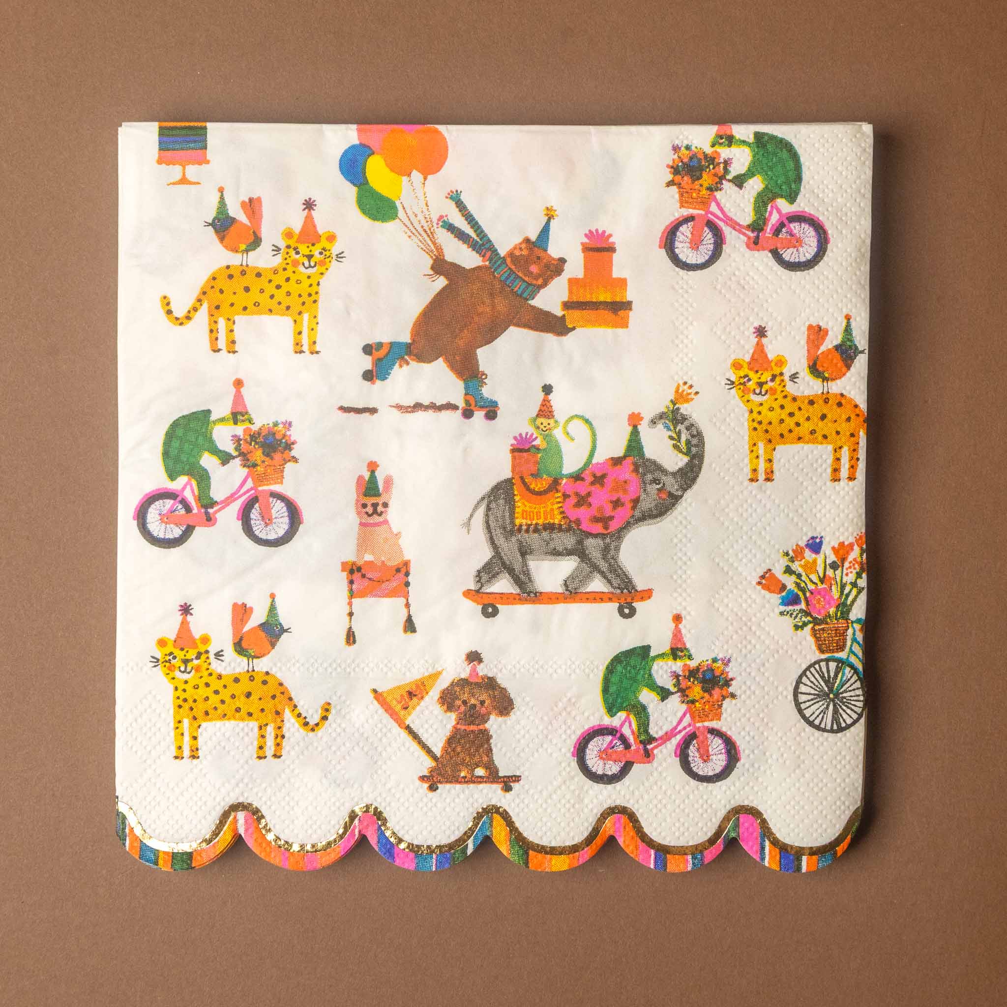 A square white paper napkin with a scalloped edge, printed with a whimsical circus-themed pattern featuring animals, bicycles, balloons, and flowers, displayed flat against a solid brown background.