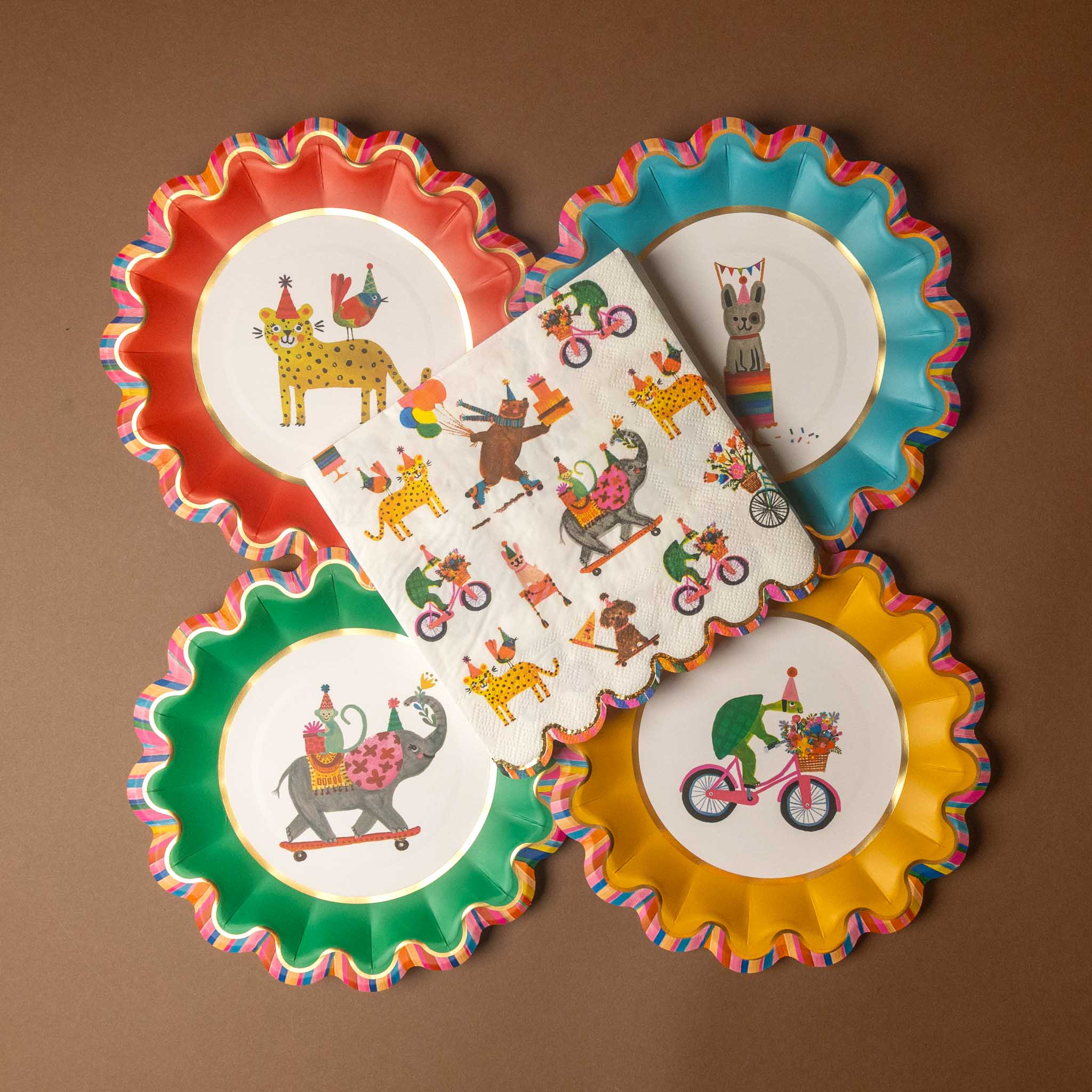 Four scalloped paper plates in red, blue, green, and yellow, arranged in a fan pattern with a matching printed napkin in the center, all on a brown background.