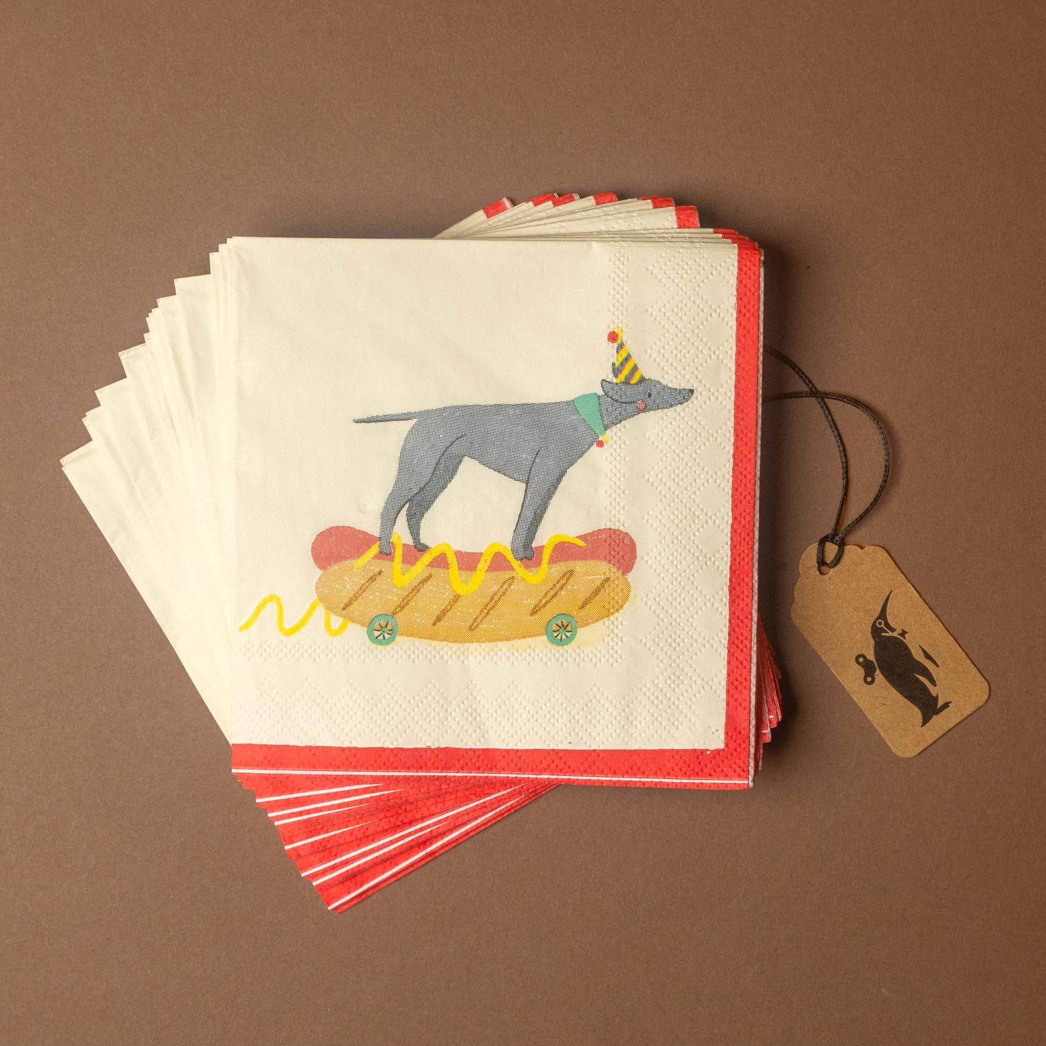 A stack of white paper napkins with red borders, printed with a cartoon gray dog standing on a hot dog with mustard, shown on a brown background with a hanging kraft paper tag.