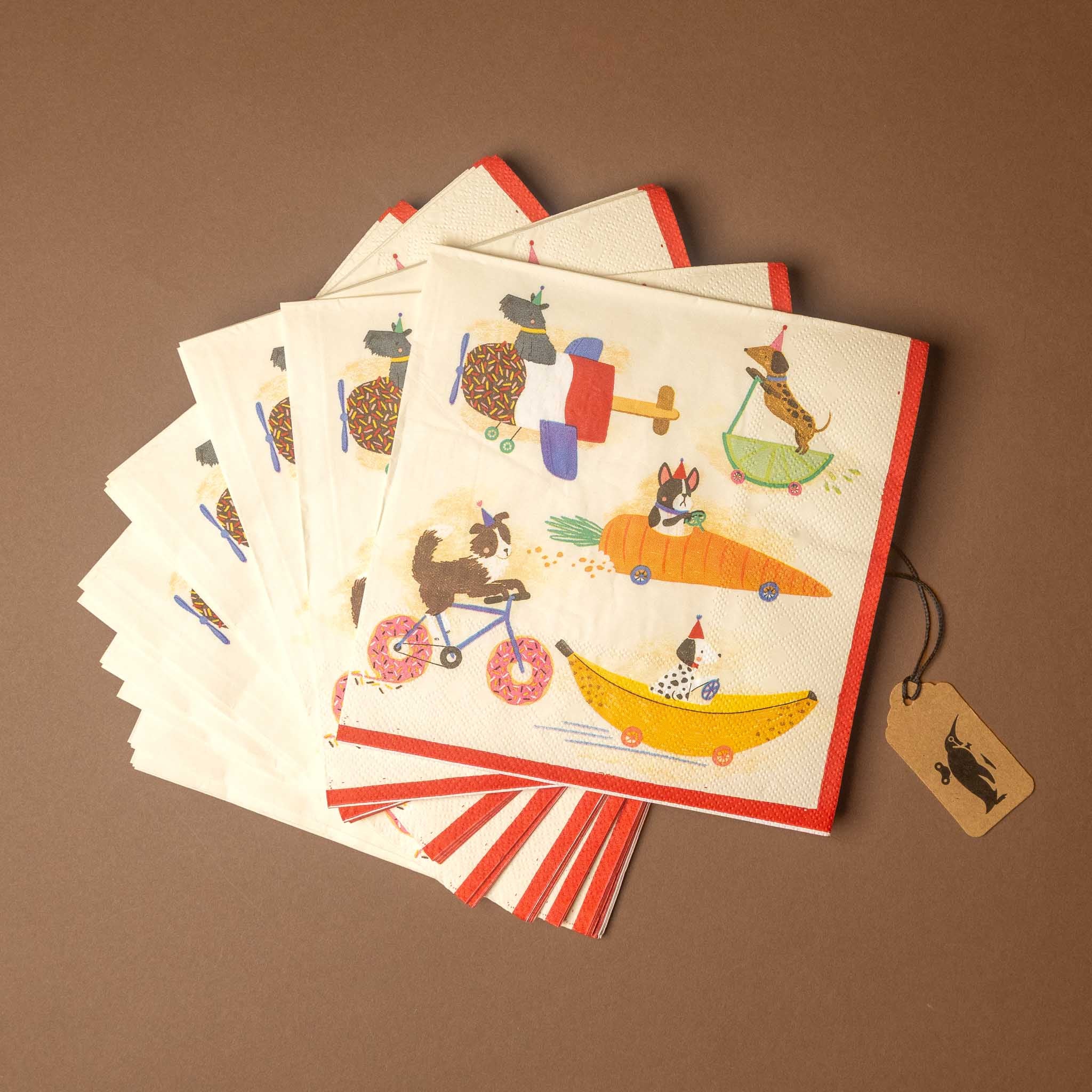 A stack of square paper napkins with red borders and a whimsical cartoon dog illustration, arranged on a brown background with a small paper tag attached.