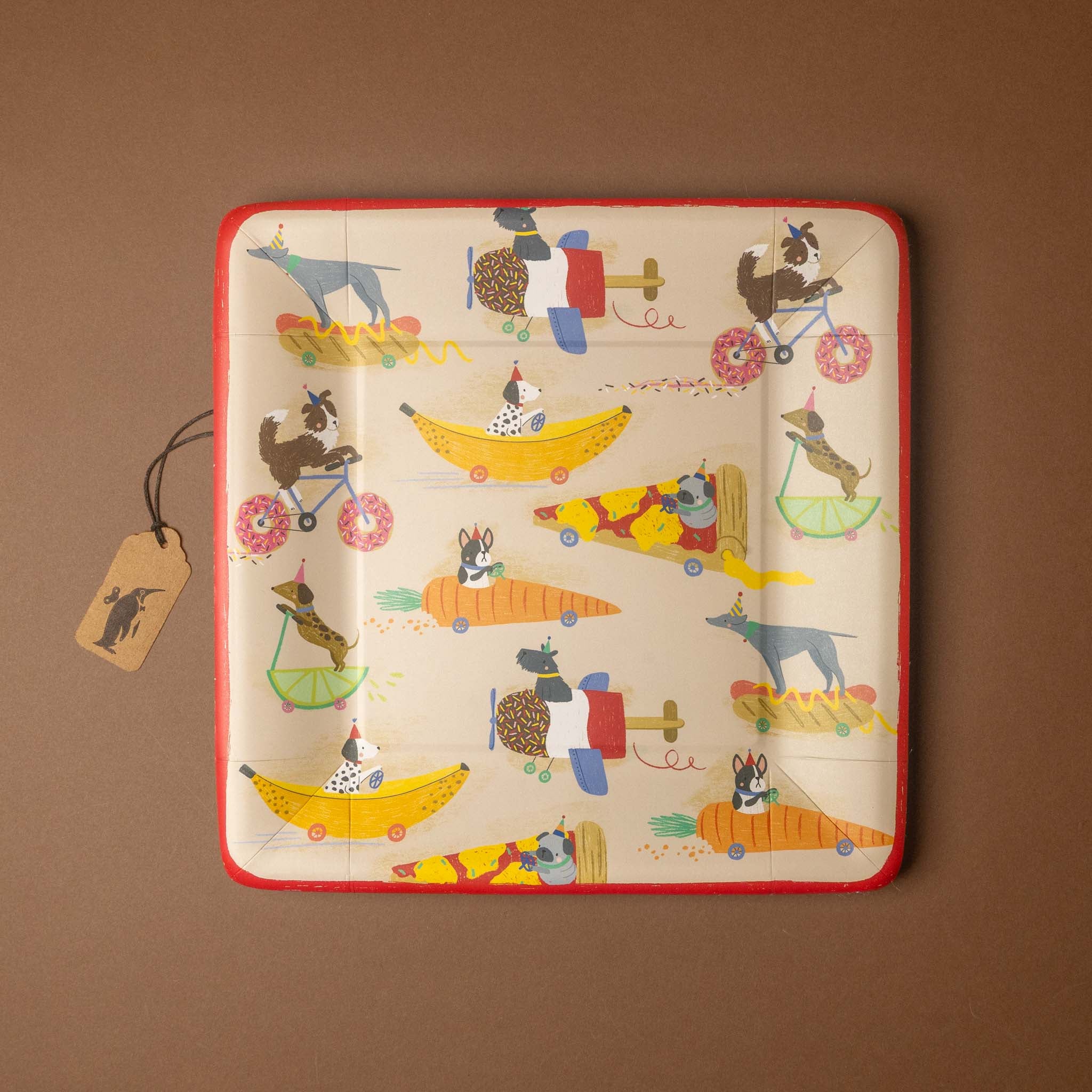 A square decorative plate with rounded corners featuring a whimsical pattern of dogs riding bicycles and flying airplanes in colorful boats. The plate has a red border and is displayed on a solid brown background with a small paper tag attached.