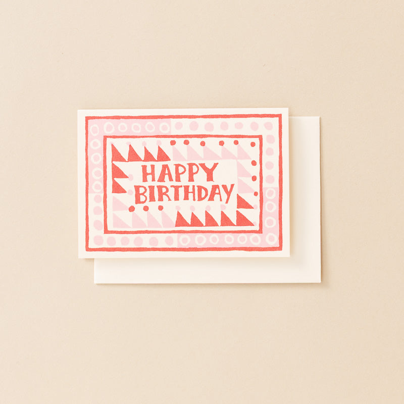 A square birthday card with a white envelope, featuring a coral red and white geometric pattern with the words 'HAPPY BIRTHDAY' in coral red text, set against a solid beige background.