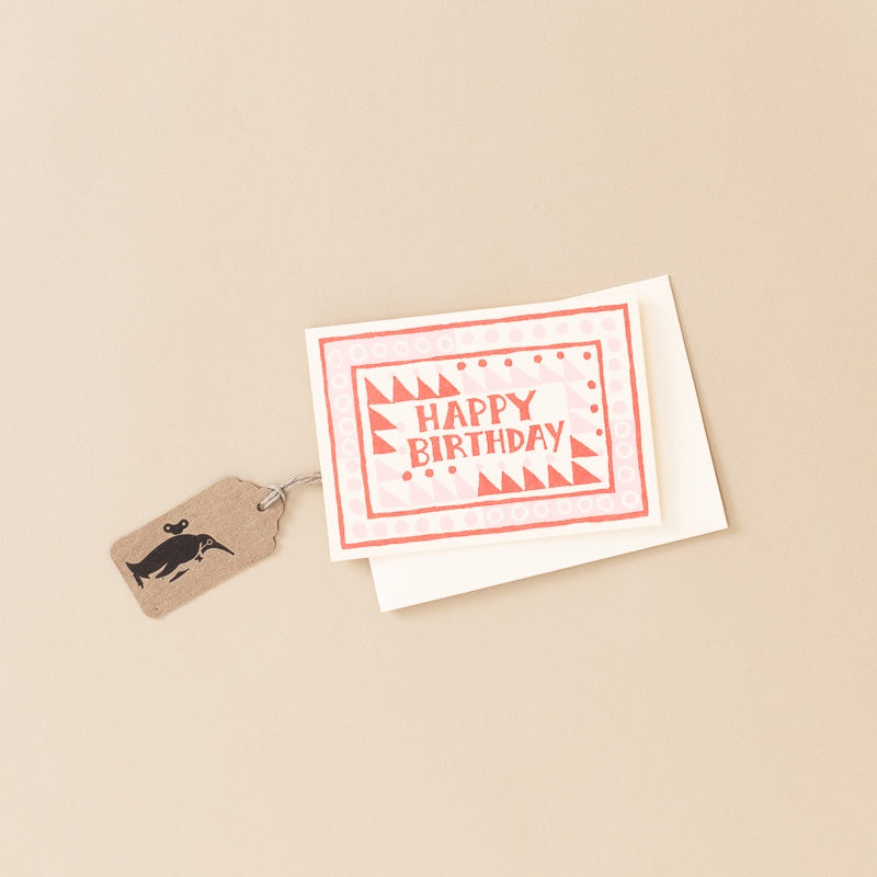 A square birthday card with a red and white patterned border and the words 'HAPPY BIRTHDAY' in red, shown with a white envelope and a small kraft paper tag with a black silhouette graphic, all on a warm beige background.