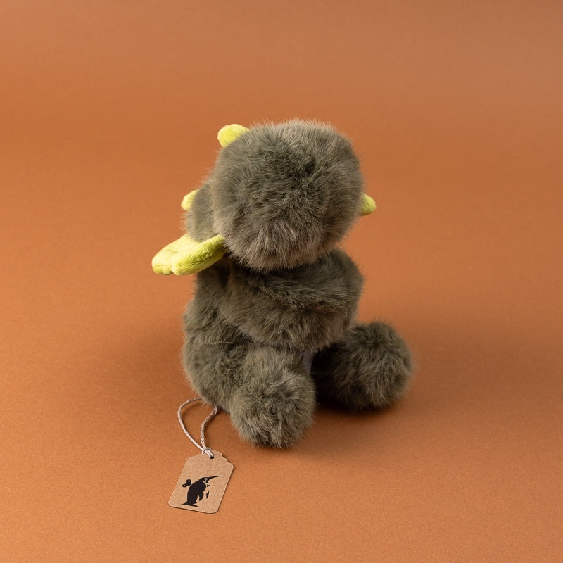 A small plush toy petite-gluepot-dino shown from the back, featuring fluffy gray-brown fur, small yellow horns, and yellow wing-like accents, sitting on a warm brown studio background with a hanging tag.