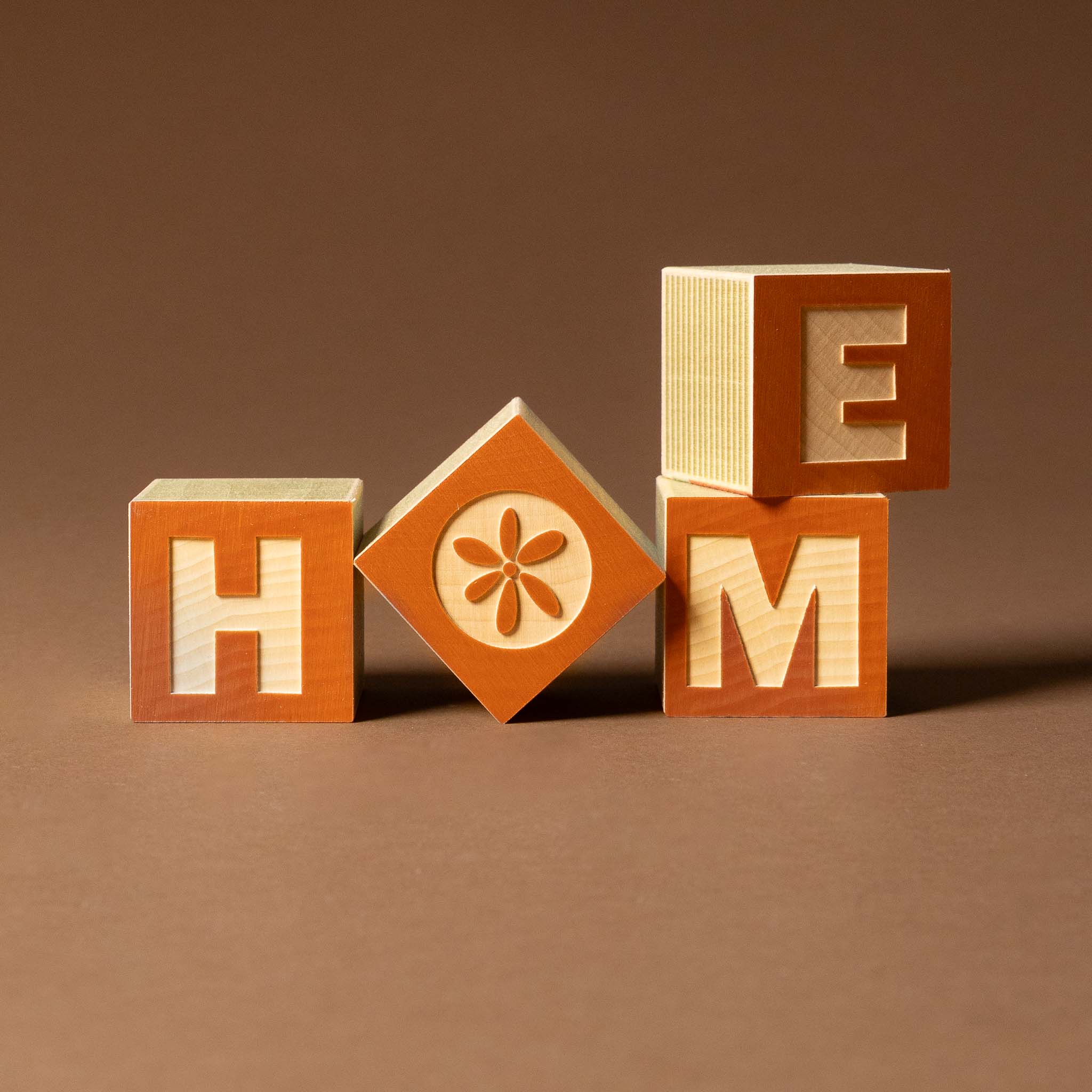 petite-wooden-gifting-block-set-home Wooden blocks spelling 'HOME' with a floral design on an orange block against a brown background