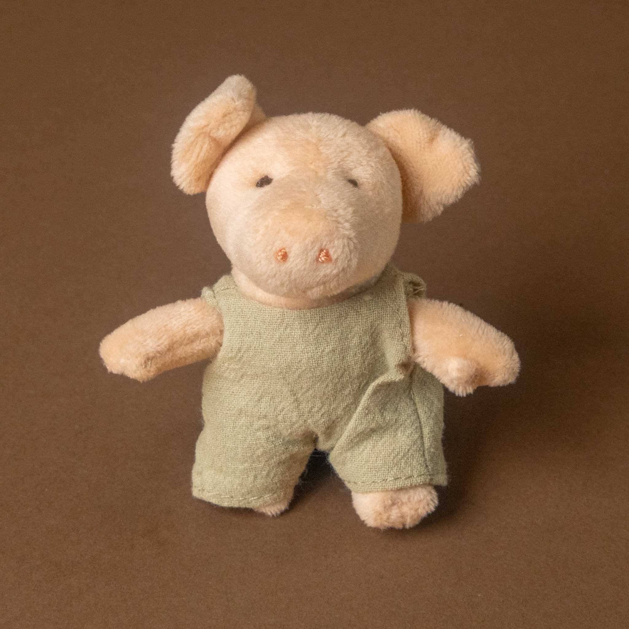 A small plush toy petits-amis-bart-pig dressed in light green overalls, photographed against a plain brown background.