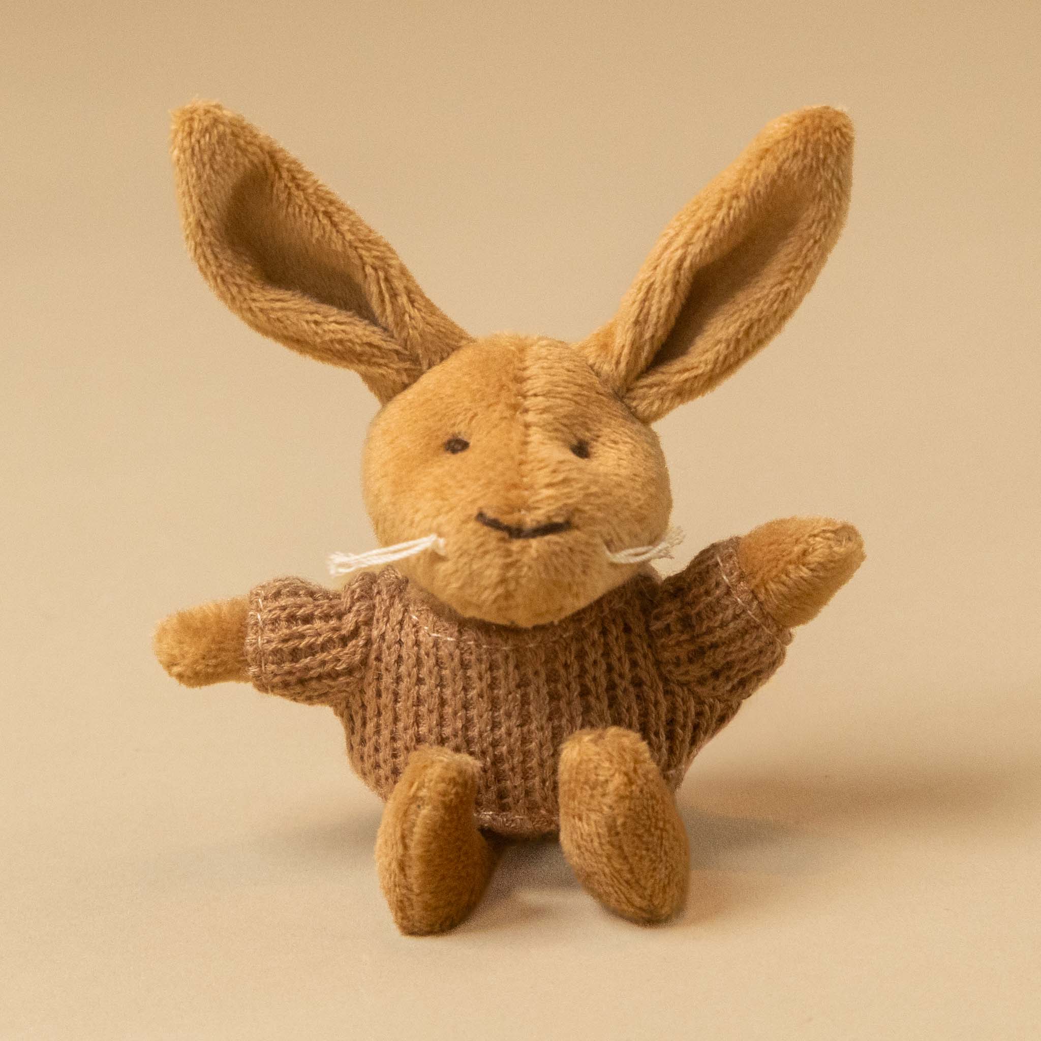 A small plushpetits-amis-lenny-rabbit with long ears, a knitted brown sweater, and white yarn whiskers, photographed against a plain warm beige background.