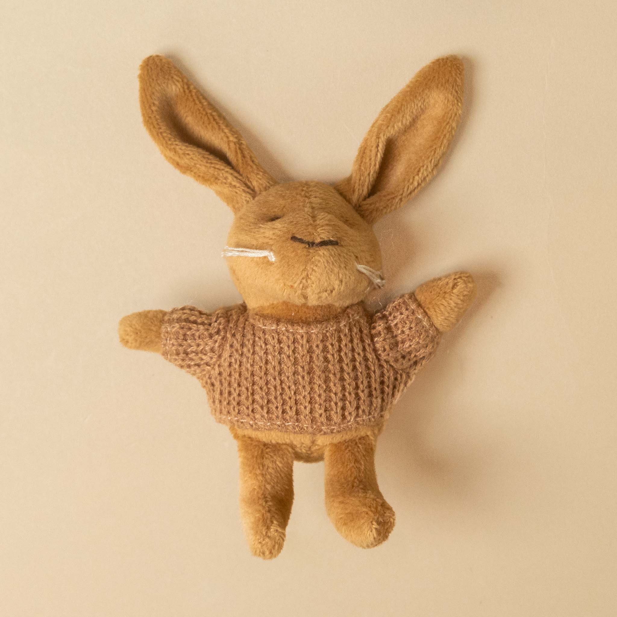 A plush stuffed petits-amis-lenny-rabbit with long ears, wearing a knitted brown sweater, photographed against a plain warm beige background.