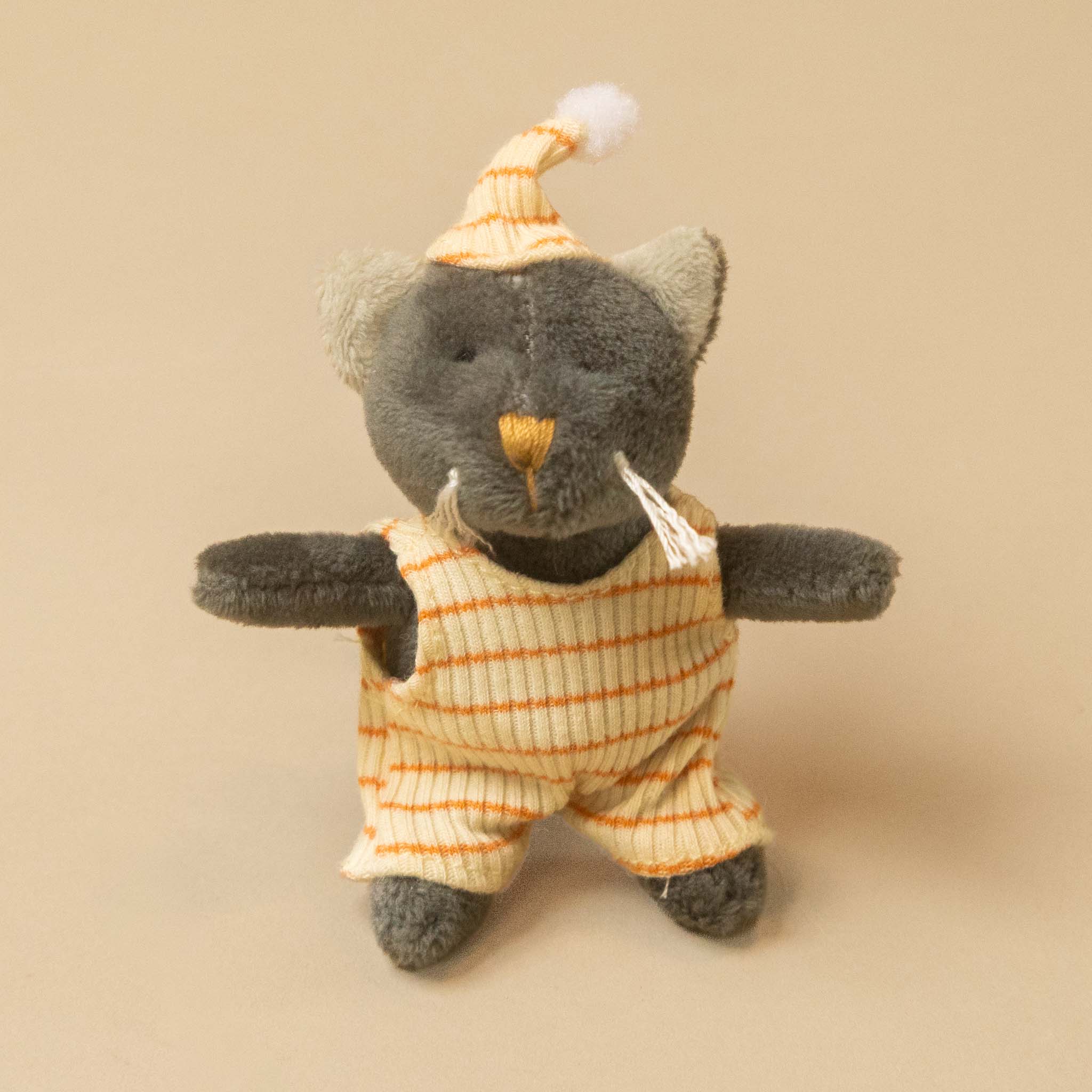 A small plush petits-amis-malo-cat wearing a cream and orange striped overalls and matching striped hat with a white pom-pom, photographed against a plain beige background.