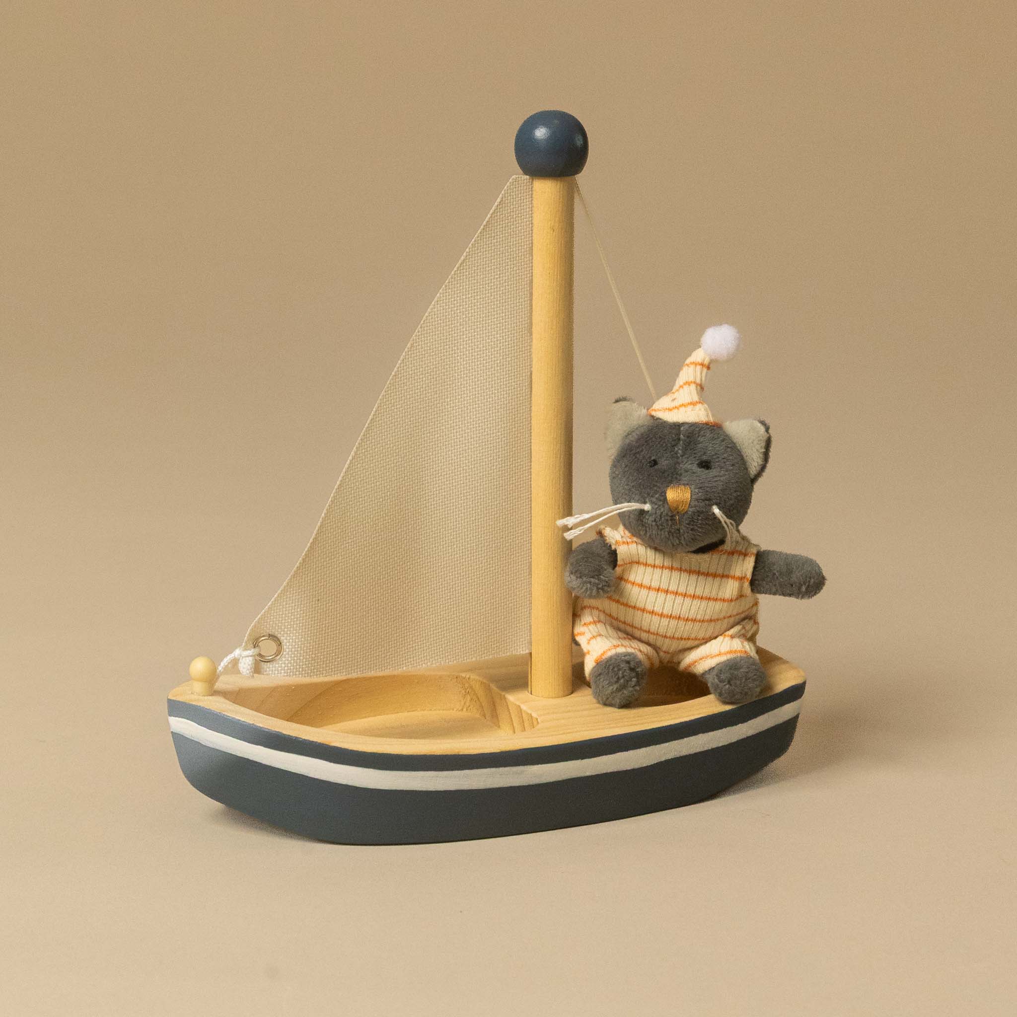 A wooden petits-amis-sail-a-way-boat-malo-cat sitting on the deck, wearing a striped orange and white outfit and a party hat. The sailboat has a natural wood mast and sail, with a dark gray hull and white stripe, set against a solid beige background.