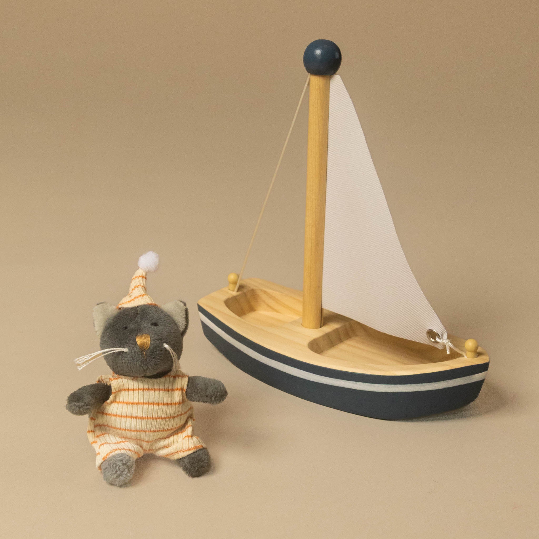 A wooden toy sailboat with a white sail and dark blue trim, displayed next to a small gray plush cat wearing orange and white striped pajamas and a matching hat, on a warm beige background.