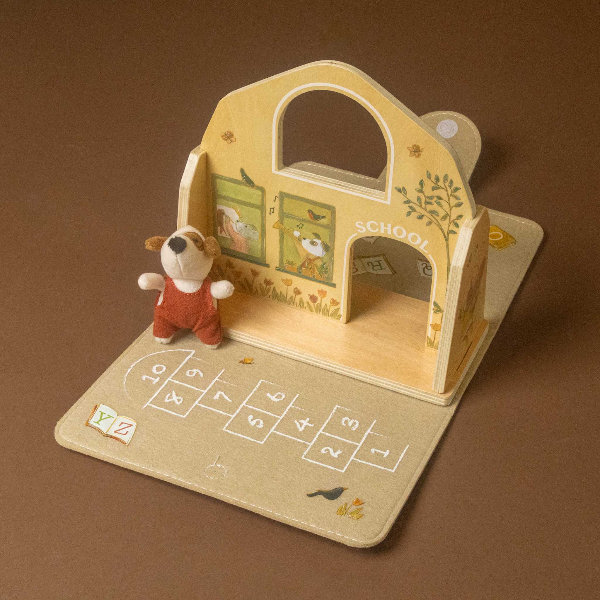 A wooden children's petits-amis-schoolhouse-play-set land hopscotch mat, shown on a plain brown background. The toy includes a small dog figurine, decorative painted panels, and printed numbers on the hopscotch base.