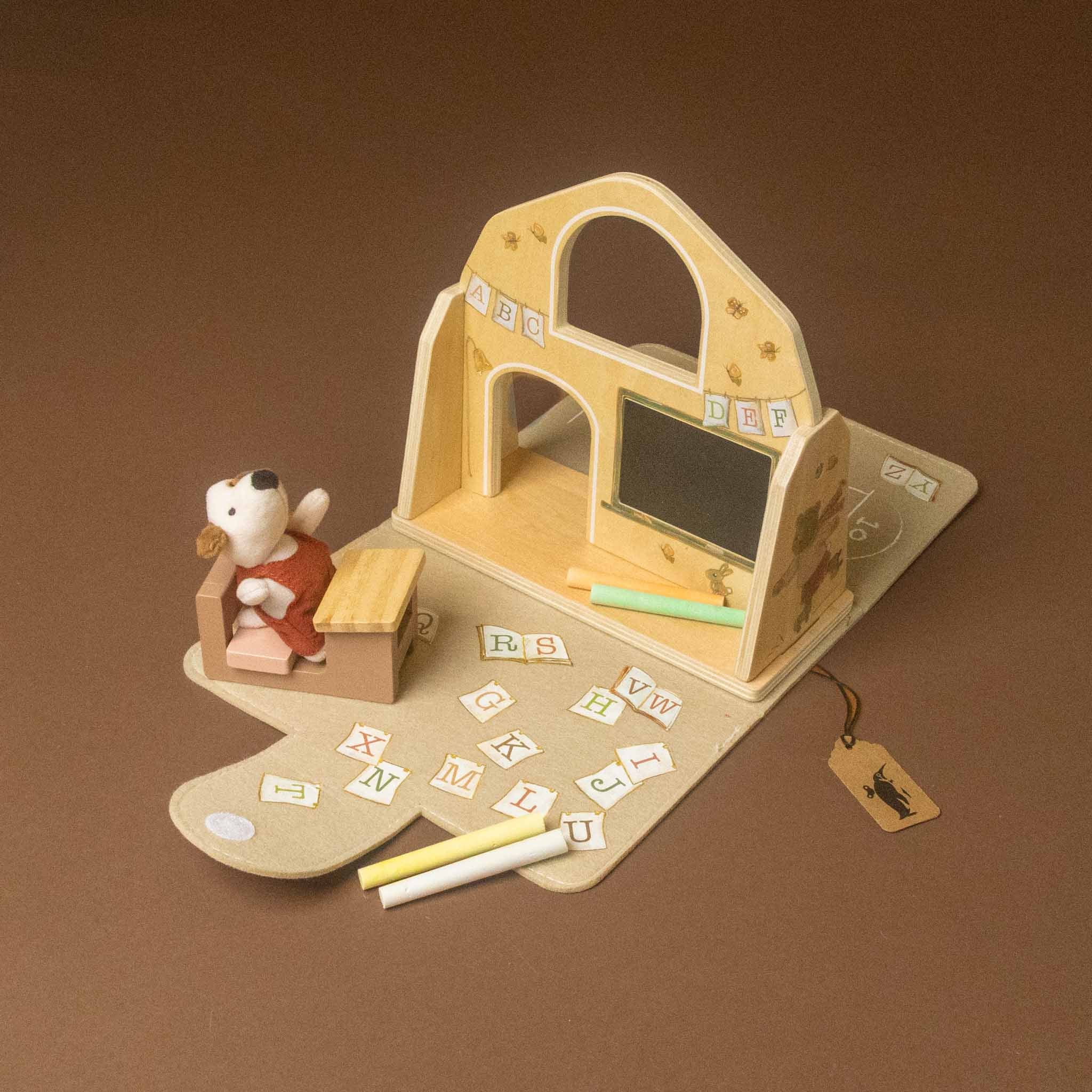 A wooden children's play school or classroom set on a brown background, featuring a small white plush dog figure in a red overalls sitting at a desk, alphabet tiles, chalk, and a hanging tag.