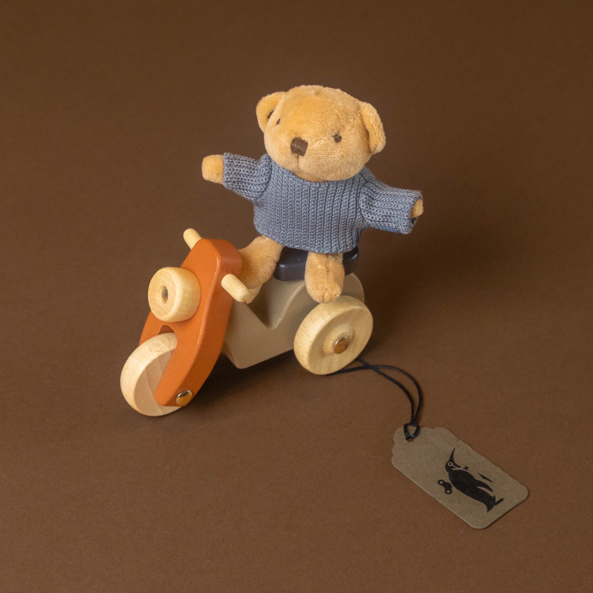 petits-amis-scooter-alexis-bear, with the bear wearing a blue knit sweater. The tricycle has an orange handlebar, beige body, and cream wheels, set against a solid brown background with a hanging tag.