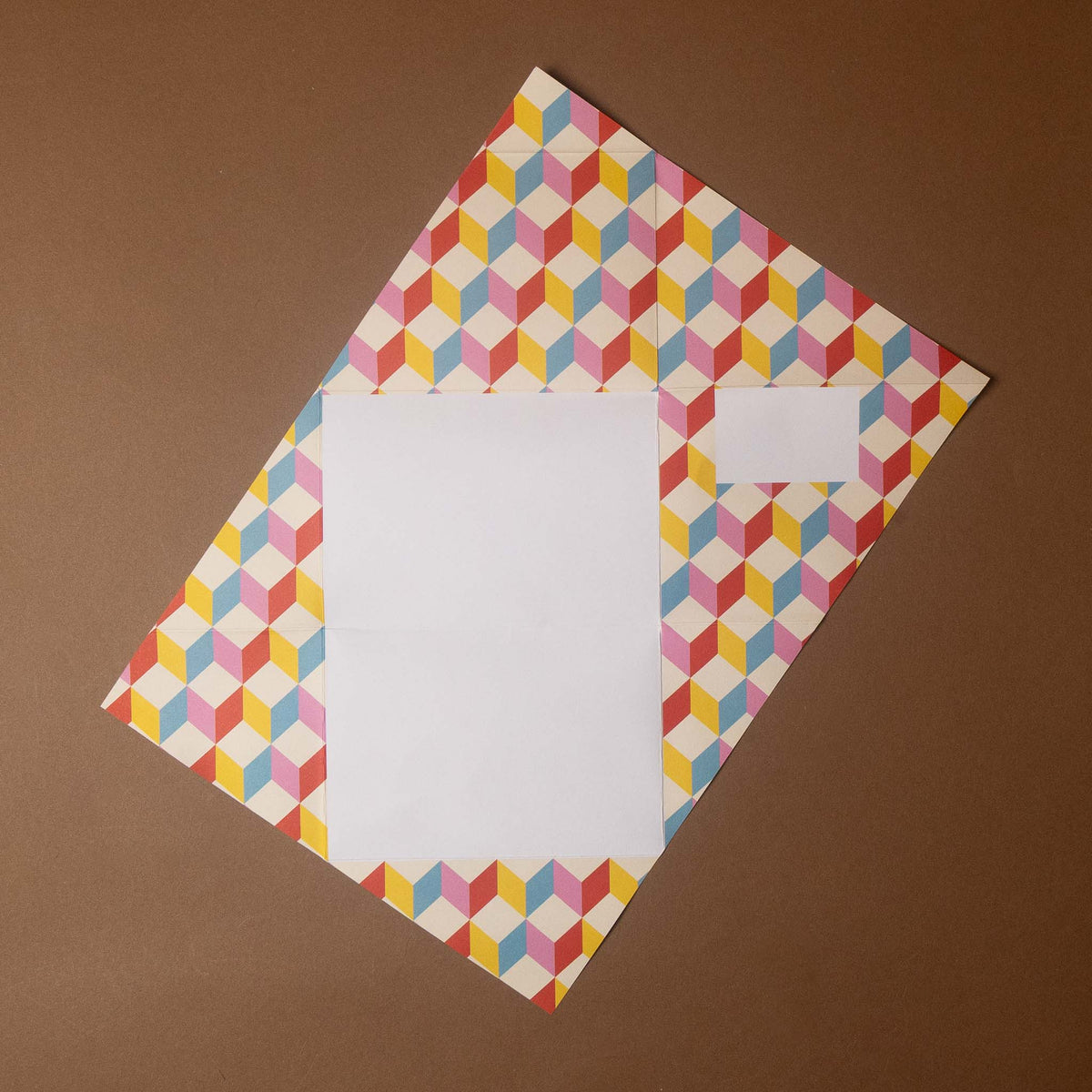 A rectangular white envelope or letterhead card with a colorful geometric pattern border, placed on a solid brown background.