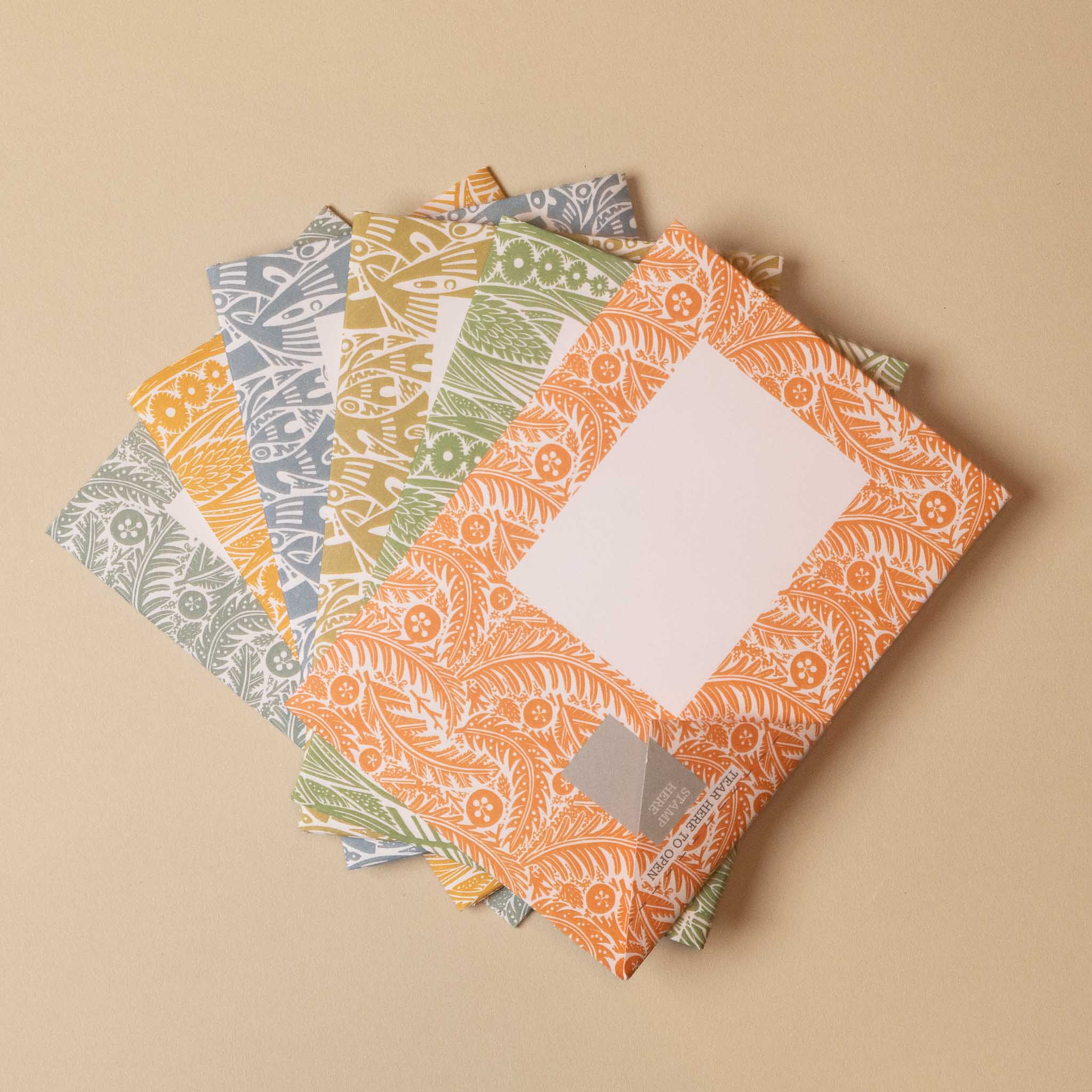A stack of patterned paper envelopes or notepads with a central blank white square, arranged on a warm beige background. The visible designs include orange, yellow, green, and blue-gray floral and leaf patterns.