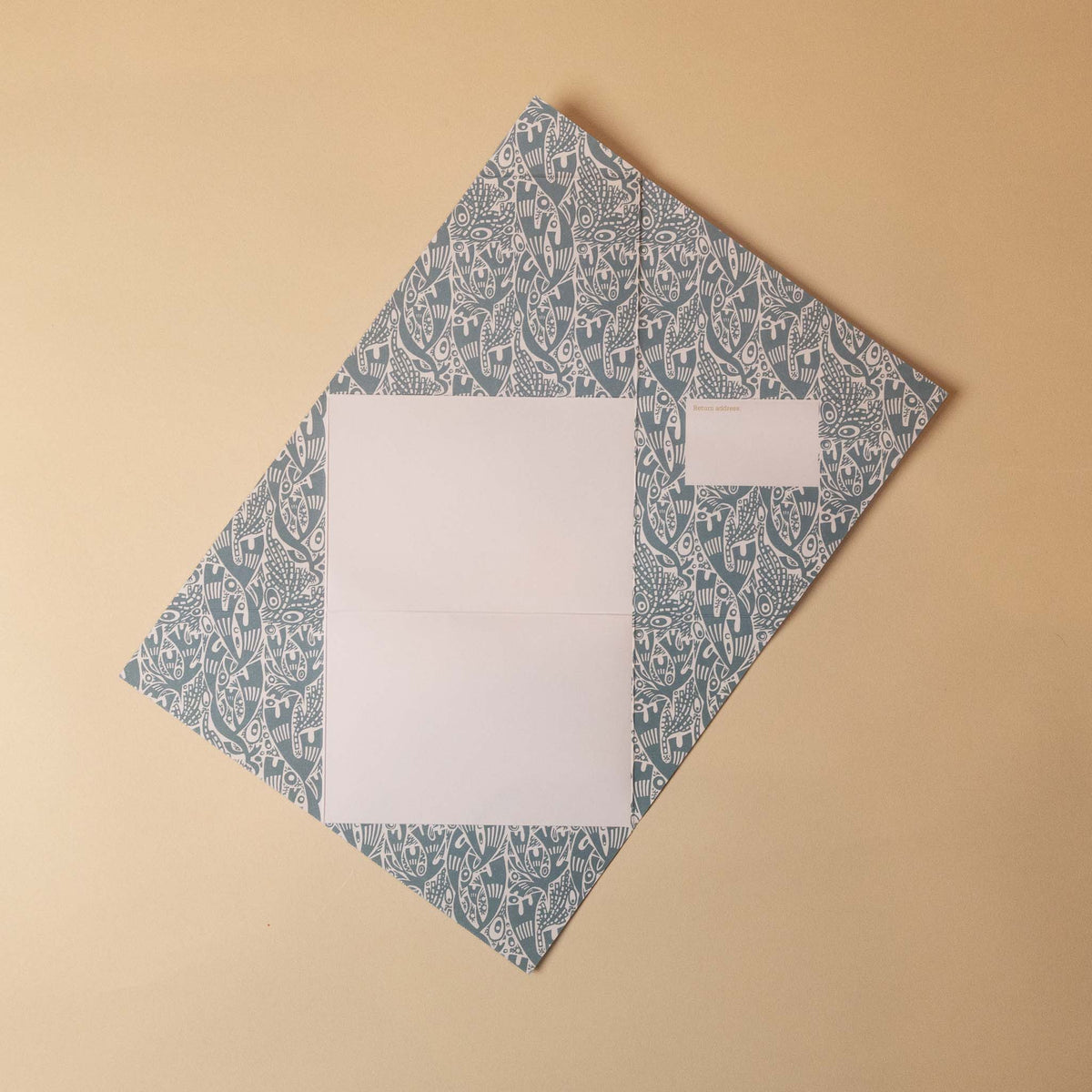 A folded paper envelope or card package with a blue and white paisley pattern, placed on a solid beige background. A white rectangular label is attached on the upper right side.