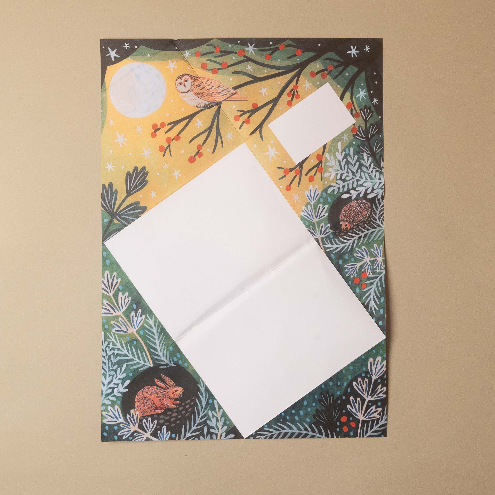 A square printed card or envelope with a winter forest illustration, laid flat on a beige background. The design shows a moon, owl, rabbit, hedgehog, berries, and pine branches, with two blank white rectangular sections overlaid near the center.