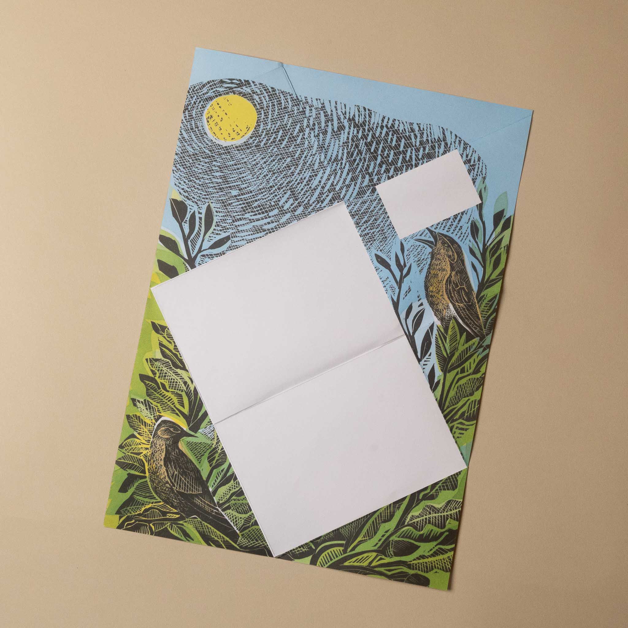 A square printed card or envelope with an illustrated bird scene, placed on a beige background. The design shows a large bird in blue-gray tones with green foliage, a yellow sun, and a small bird near the lower right.