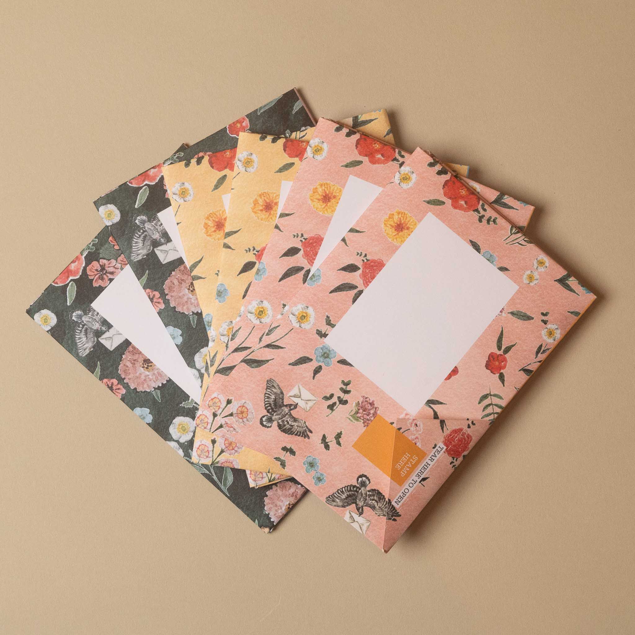 Four floral patterned envelopes fanned out on a warm beige background. The envelopes feature different colorways: a dark gray/black with white flowers, a soft yellow with orange flowers, a dusty pink with red flowers and butterflies, and a peach-pink with orange flowers and butterflies.