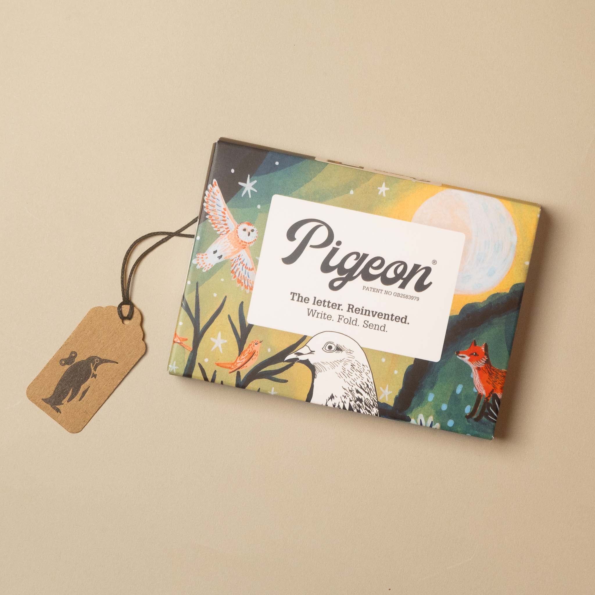 A rectangular Pigeon Envelope & Company gift box with a colorful illustrated cover, shown on a warm beige background with a small kraft paper tag attached by a string.