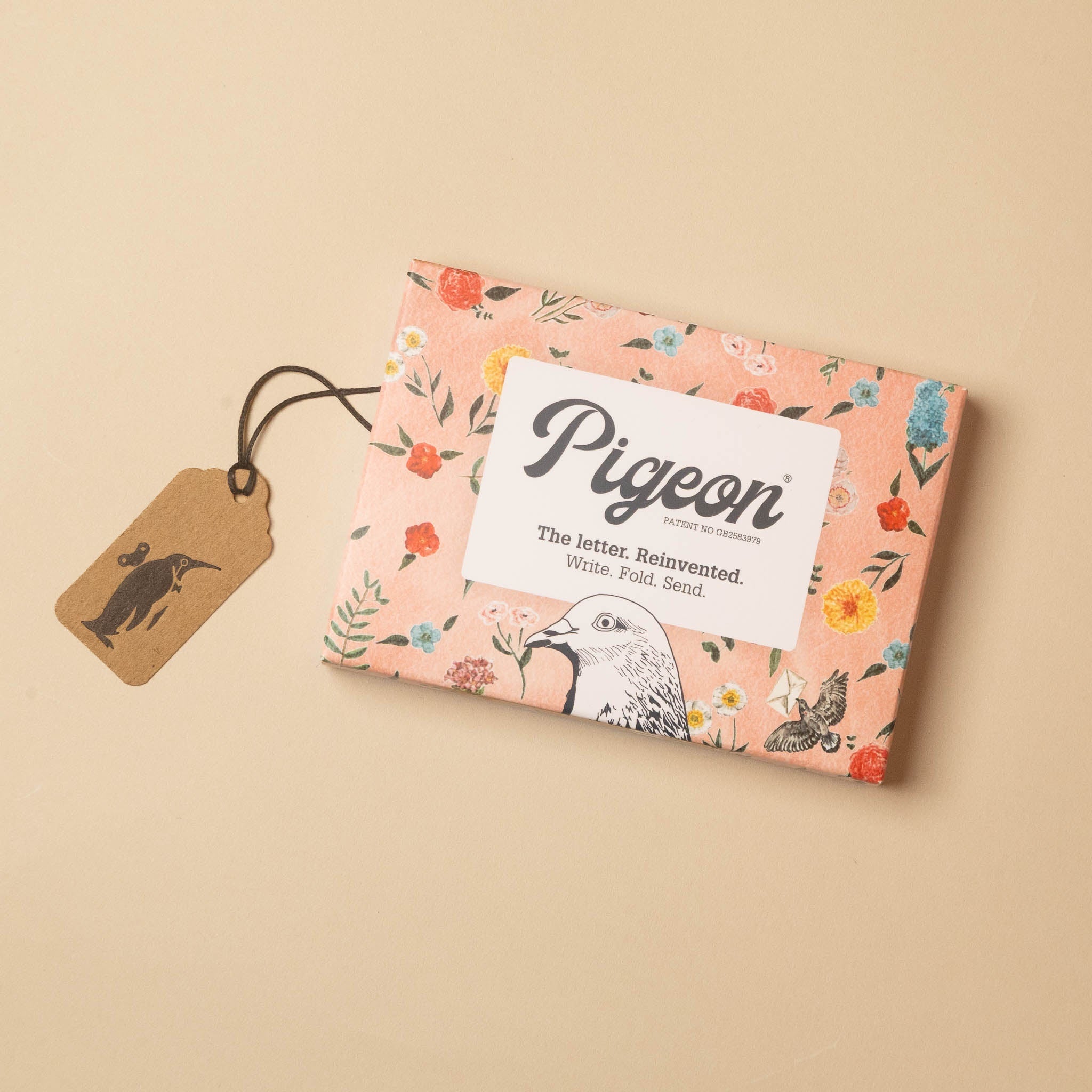 A rectangular boxed product wrapped in peach floral paper with a white label reading 'Pigeon' and a small brown paper tag with a black bird silhouette, photographed on a warm beige background.
