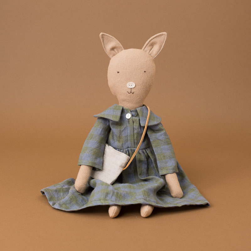 A plush stuffed pig doll shaped like a  wearing a blue and green plaid dress with a white pocket and a small tan crossbody bag, posed against a solid warm brown background.
