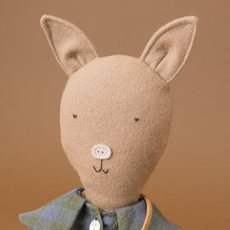 A handmade felt stuffed pig, with long upright ears, simple embroidered facial features, and a blue-green plaid collar, shown against a warm tan background.