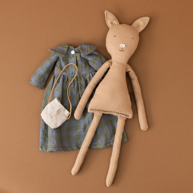 A handmade felt toy pig in a tan color, styled next to a blue-green plaid doll dress and a small cream fabric purse, all laid flat on a solid brown background.