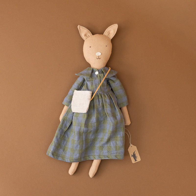A plush stuffed pig doll shaped like a  wearing a blue and green plaid dress with a white pocket and a small tan crossbody bag, posed against a solid warm brown background.