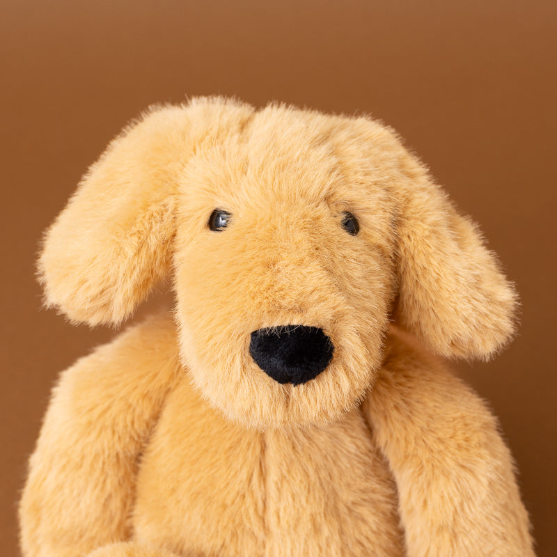 A close-up of a plush dog toy with soft tan fur, black eyes, and a black nose against a solid brown background.