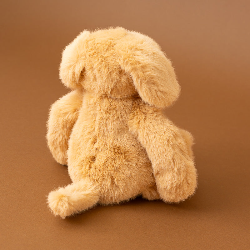 A light tan plush stuffed bunny shown from the back, sitting on a solid brown studio background.