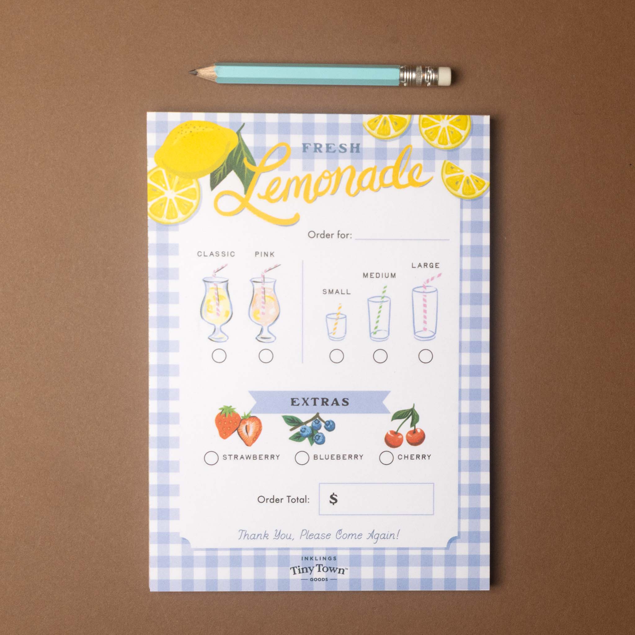 A printable lemonade order card or sign with illustrated lemon graphics, drink size options, and extra flavor checkboxes, photographed flat on a brown surface with a teal pencil above it.