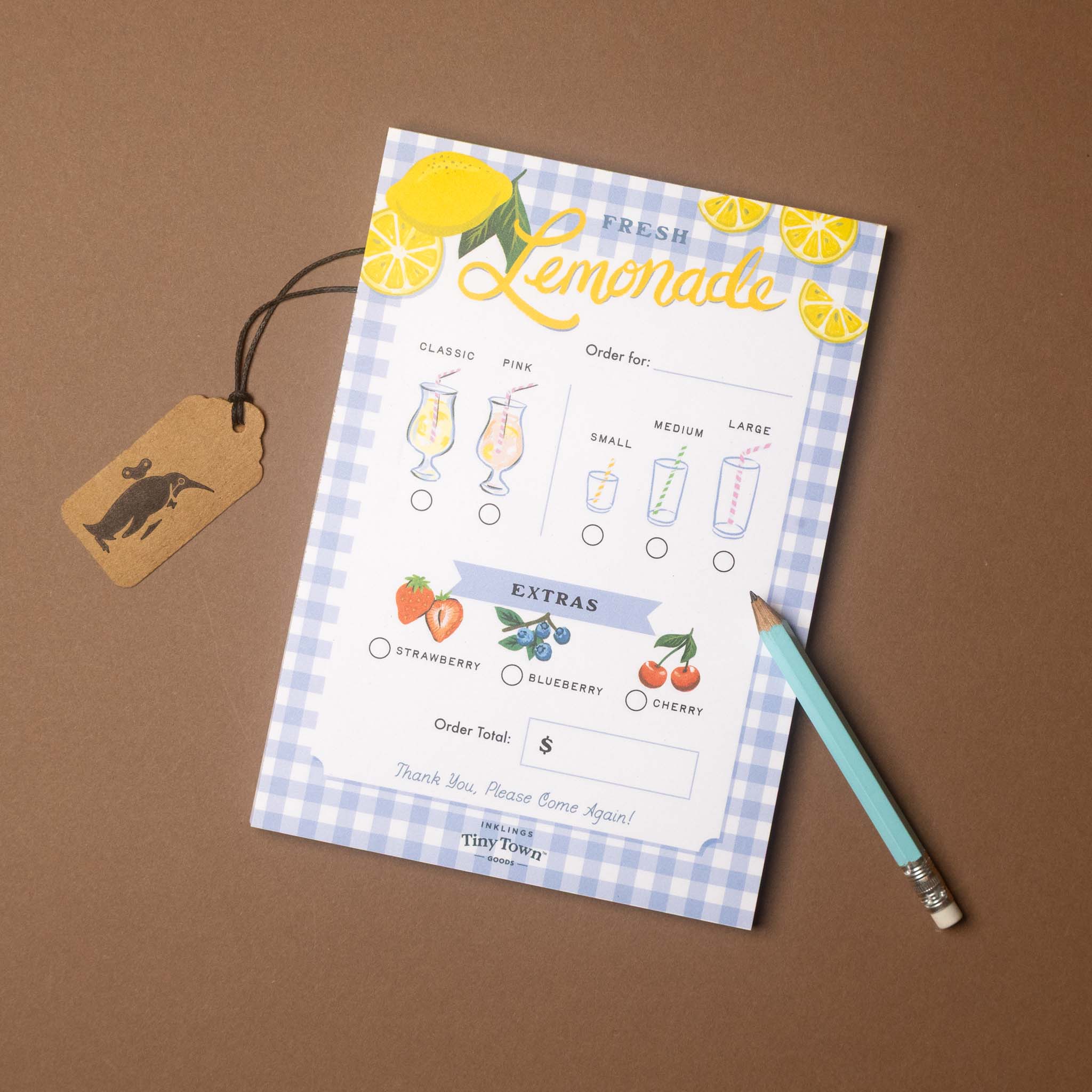 A printed lemonade order card with lemon illustrations, glass size guides, and an extras section, styled on a brown surface with a hanging tag and a pencil.