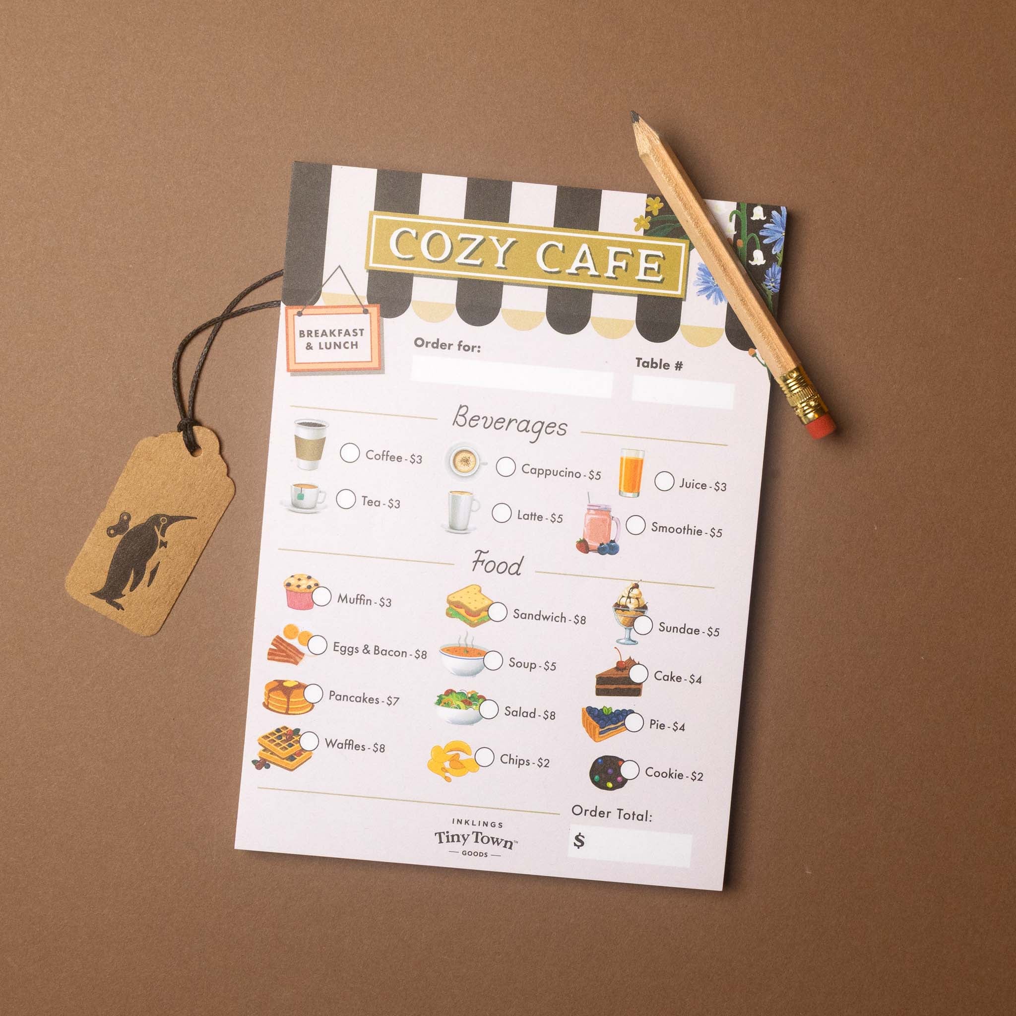 A printed cafe order card titled 'COZY CAFE' laid on a brown surface, with a pencil resting on top and a small hanging tag attached by a string. The card includes sections for beverages, food, table number, and a price field, with illustrated menu items and small logo graphics.