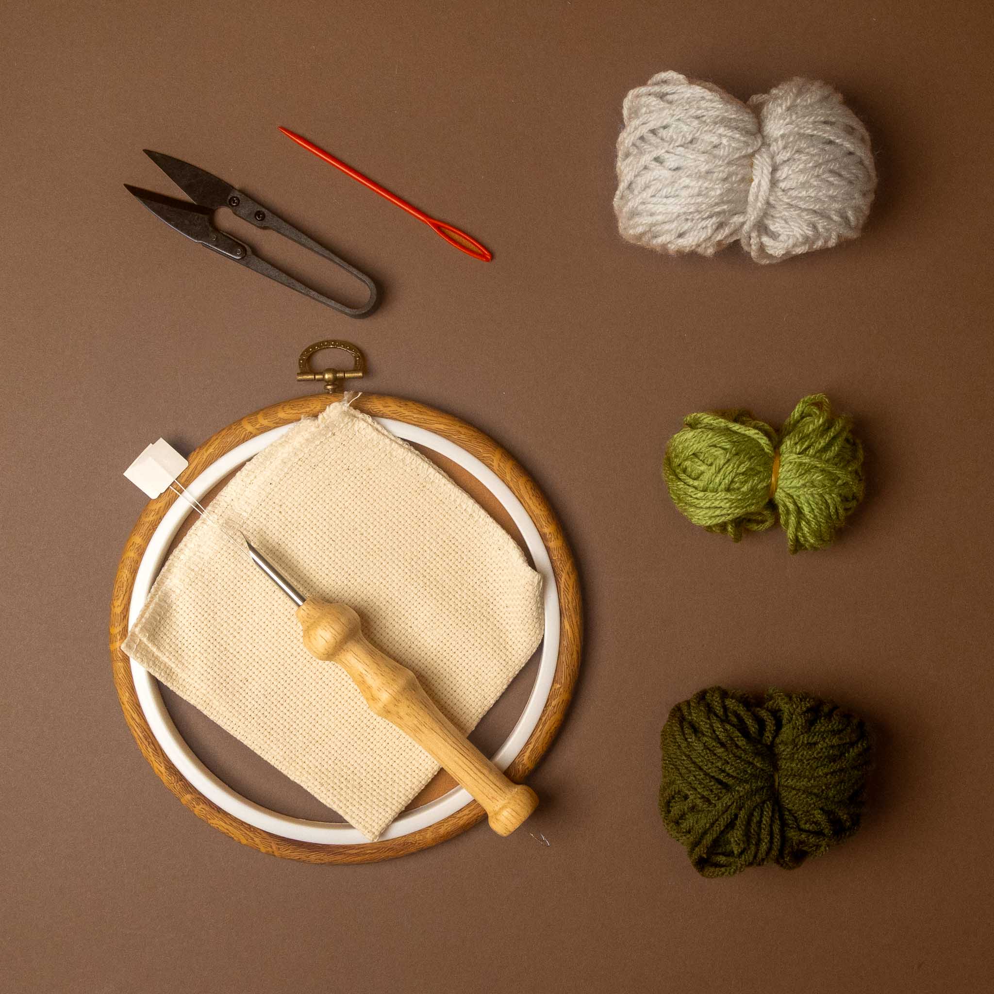 A flat-lay of knitting or embroidery supplies on a brown background, including a wooden hoop with cream fabric, a wooden needle, a red crochet hook, metal scissors, and three balls of yarn in off-white, olive green, and dark green.