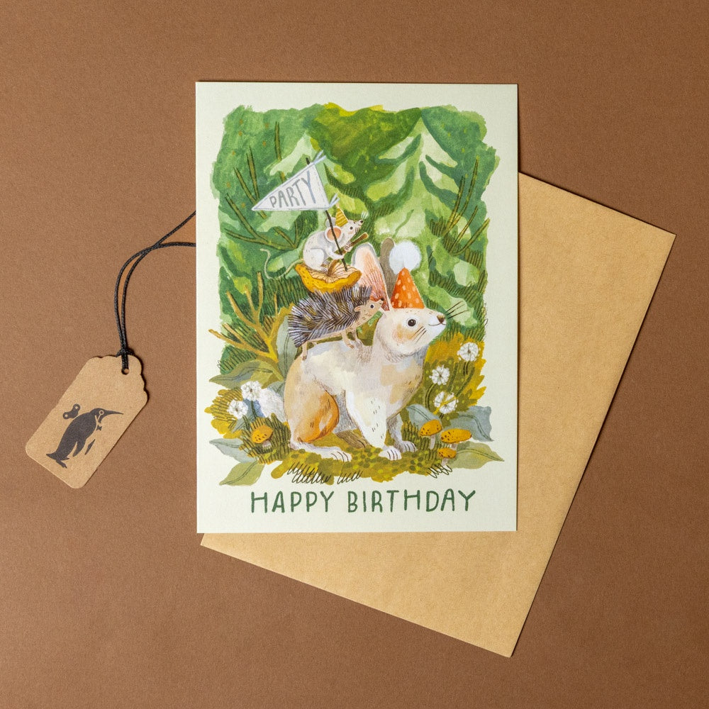 rabbit-celebration-greeting-card with birthday-forest-themed illustration of animals and a party hat, accompanied by a tag and envelope on a brown background.
