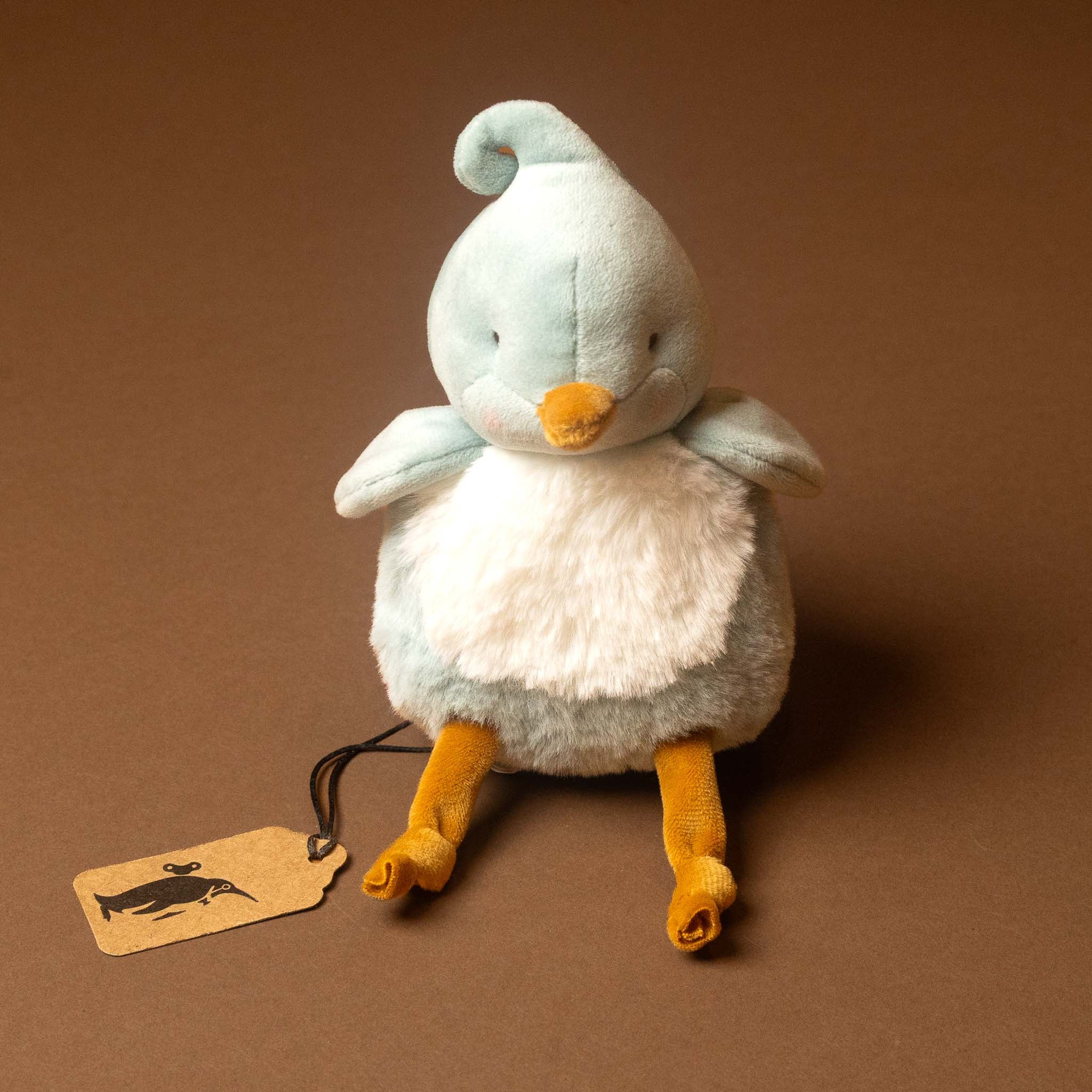 A plush roly-poly-birdie-tweet with pale blue feathers, a white belly, and orange beak and legs, sitting on a brown background with a small paper tag attached.