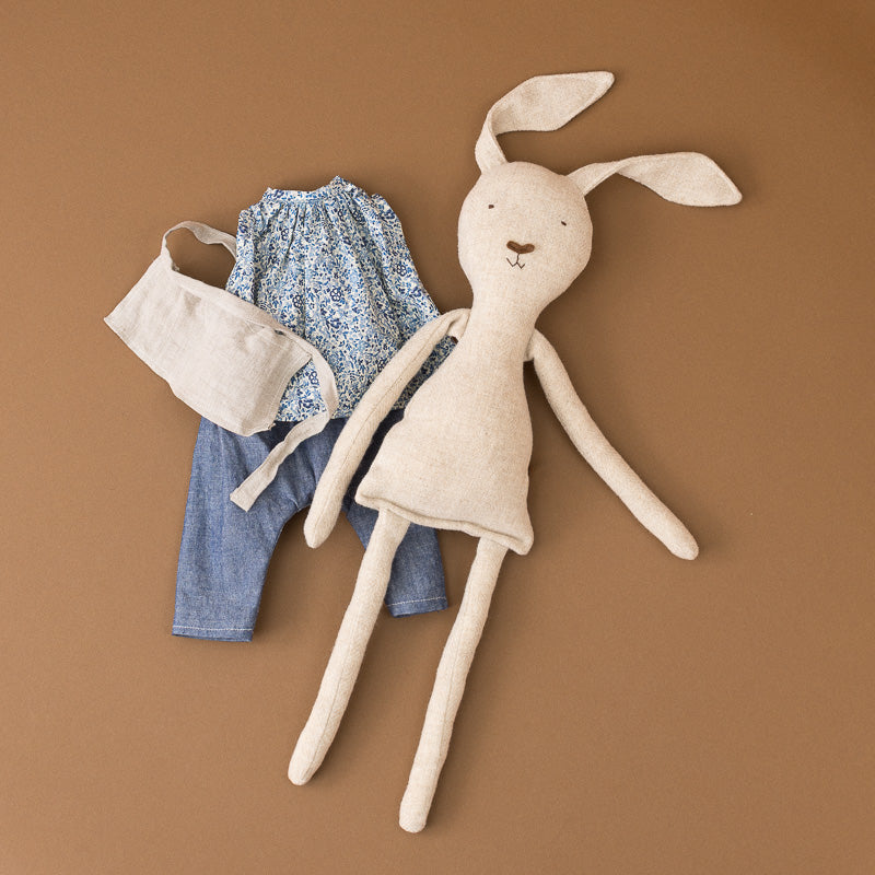 A handmade plush bunny doll with long ears and limbs, styled with a blue floral top, denim pants, and a cream fabric bag on a solid brown background.