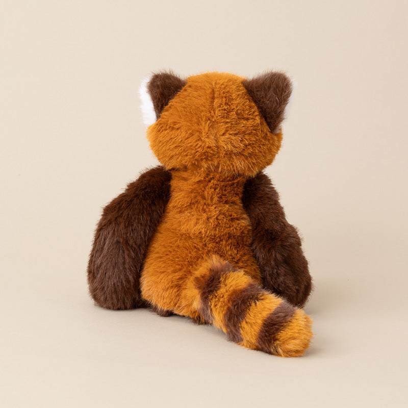 A plush red panda toy shown from the back, featuring orange-brown fur, dark brown arms and ears, and a striped tail, photographed against a solid beige background.