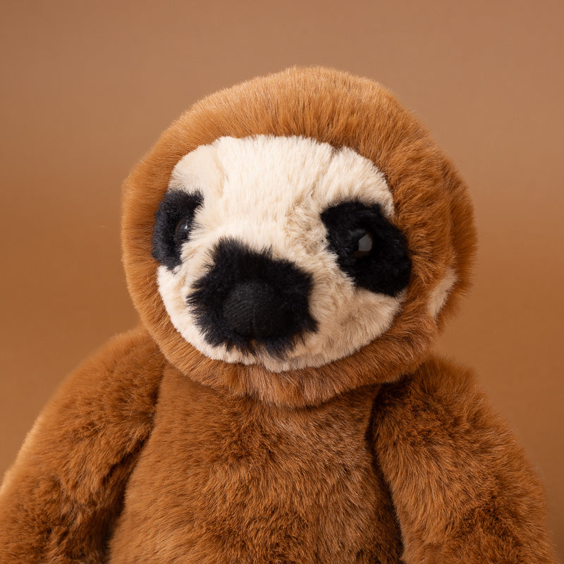 A close-up of a plush sloth toy with brown fur, a cream-colored face, and black facial markings, set against a solid warm brown background.