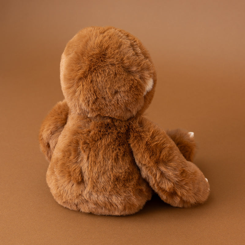 A brown plush teddy bear viewed from the back, sitting on a smooth brown studio background.