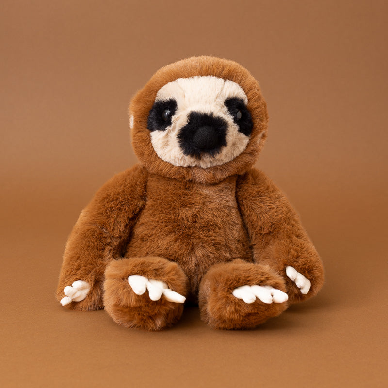 A plush sloth toy sitting upright against a solid warm brown background. The sloth has medium brown fur, a cream-colored face, black eyes and nose, and white claws.