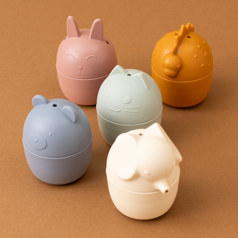 Five small egg-shaped animal-shaped containers in pastel and warm tones, arranged on a plain brown background. The animals include bunny, cat, bear, dinosaur, and elephant faces.